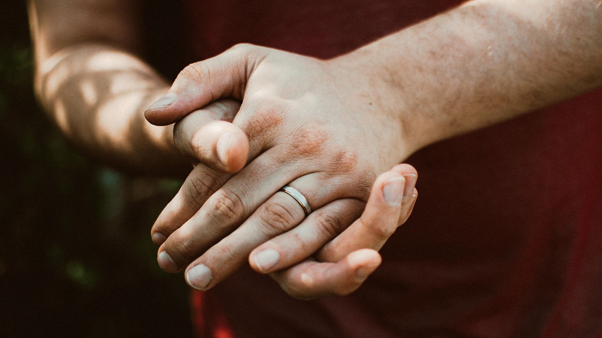 A close-up shot of clasped hands with a wedding ring, captured outdoors in natural light.