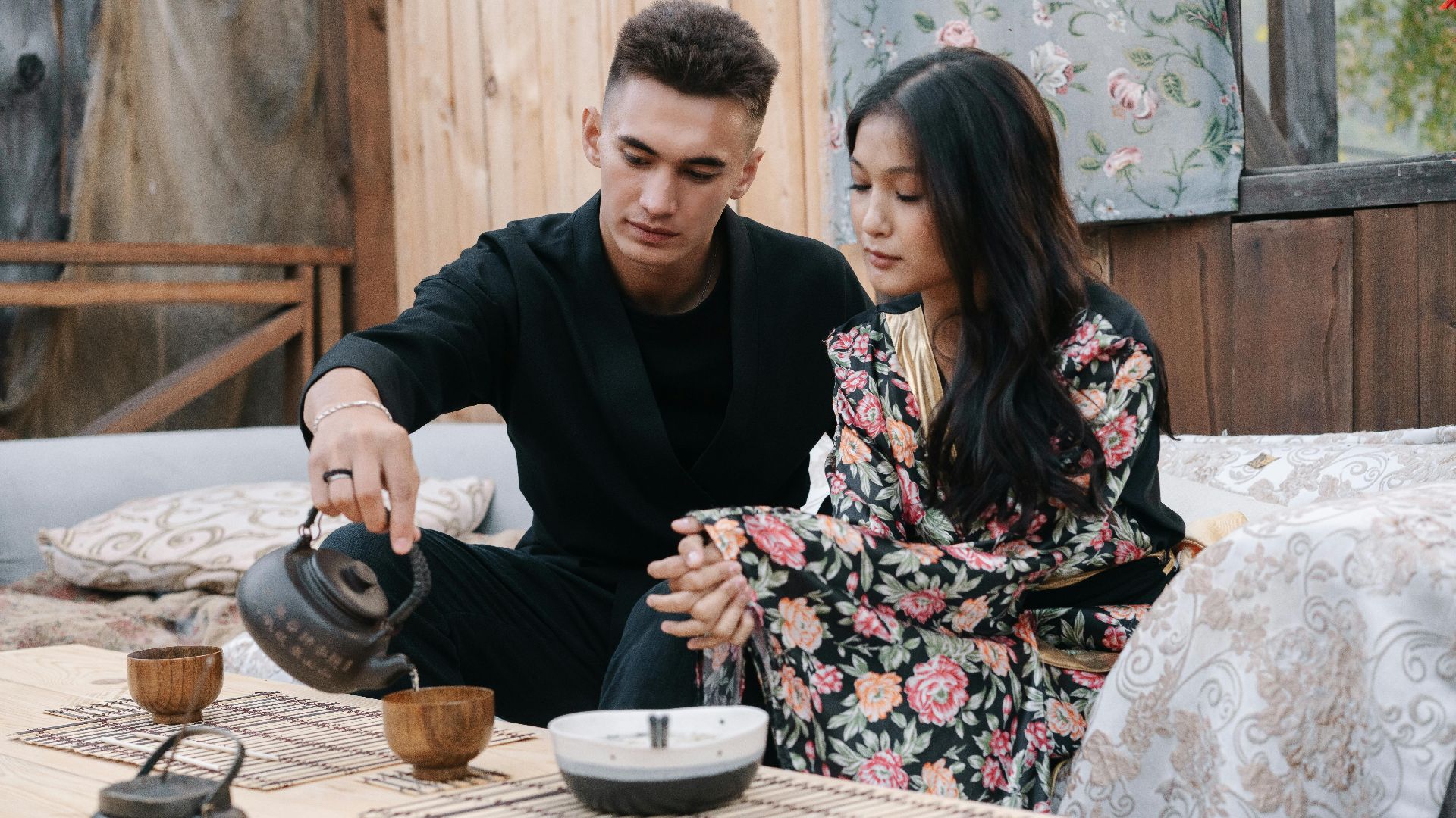 A young couple shares a quiet moment over a traditional tea ceremony indoors, surrounded by cozy decor.
