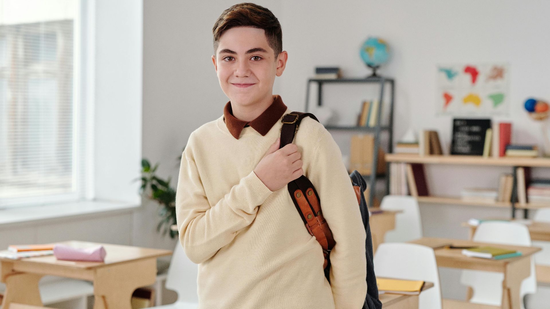 A smiling schoolboy confidently standing in a modern classroom with his backpack.