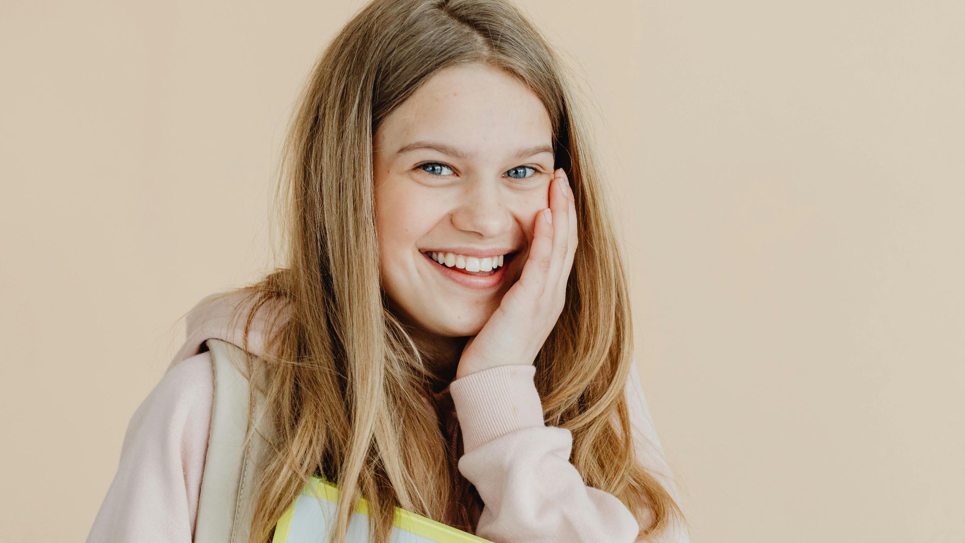 Bright studio shot of a smiling teenage girl with blonde hair holding a book. Casual and joyful mood.