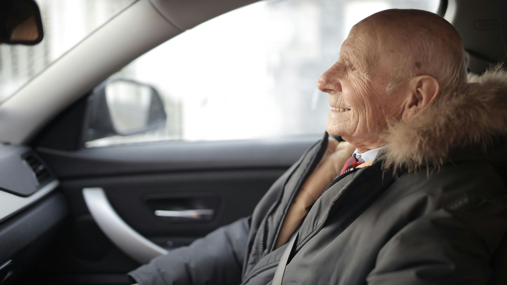 Side view of content elderly male in suit and outerwear sitting in front seat of contemporary automobile and looking away
