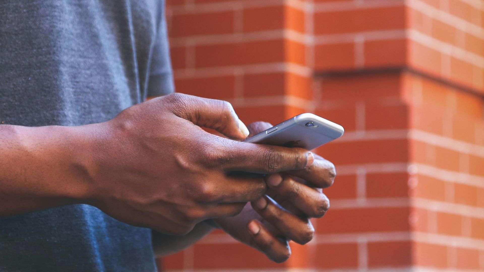 A smiling man checks his smartphone while leaning against a brick wall outdoors.