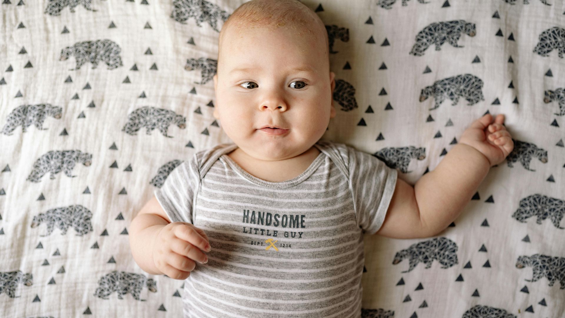 Cute baby boy wearing a striped onesie, lying on a bear-patterned blanket with a smile.