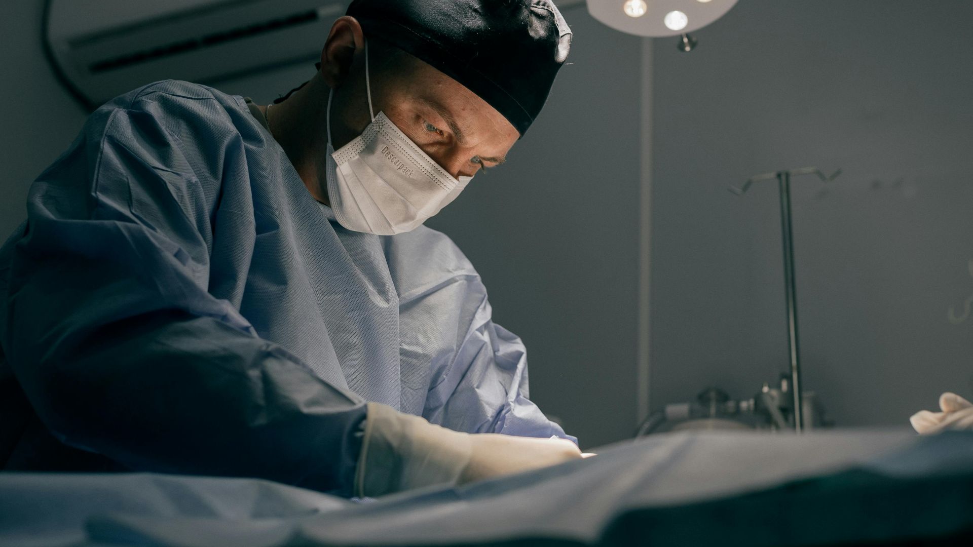 A surgeon in an operating room performing a procedure under surgical lights, wearing mask and scrubs.