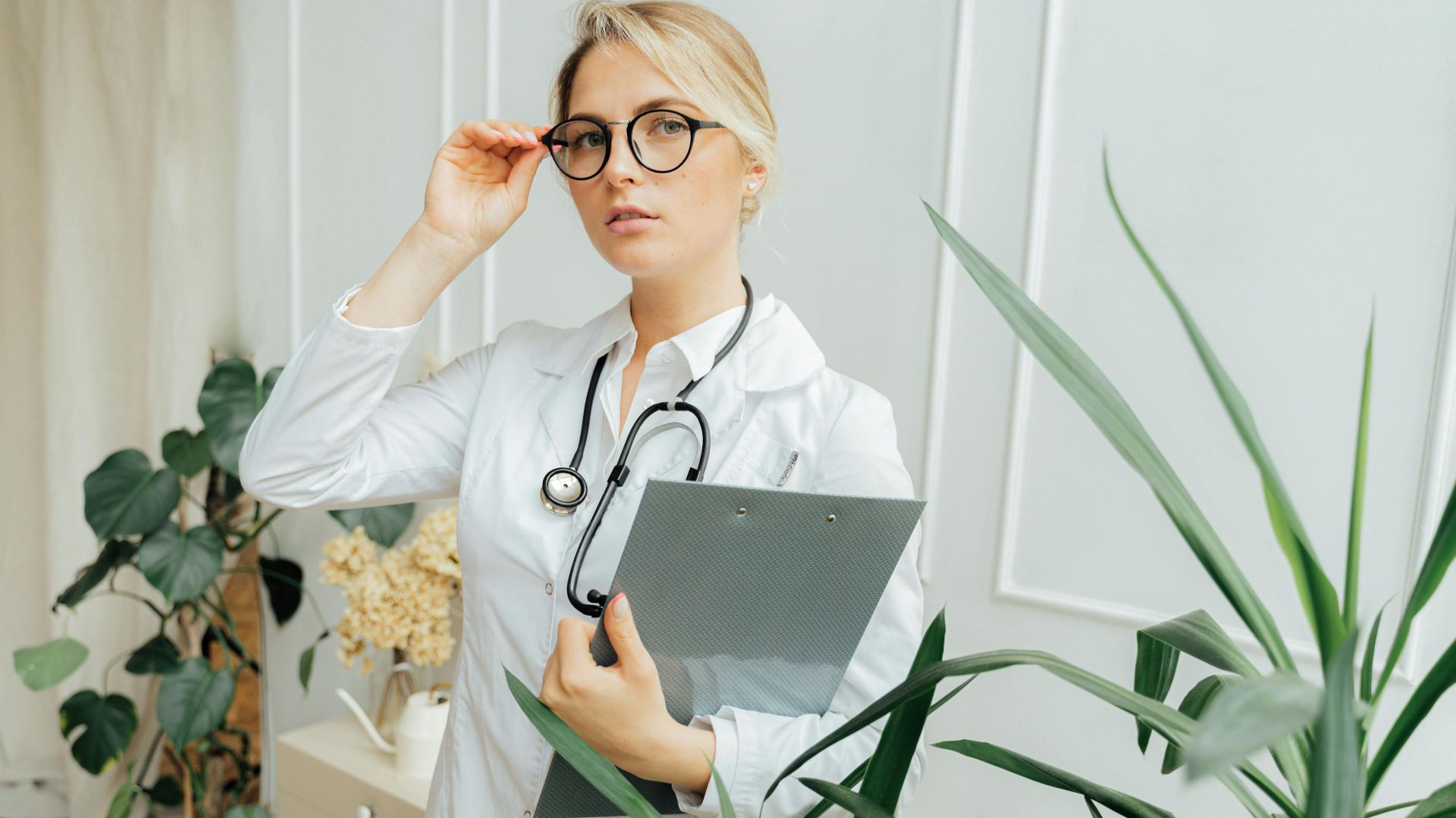 Confident female doctor wearing eyeglasses and holding a clipboard in an indoor office setting.