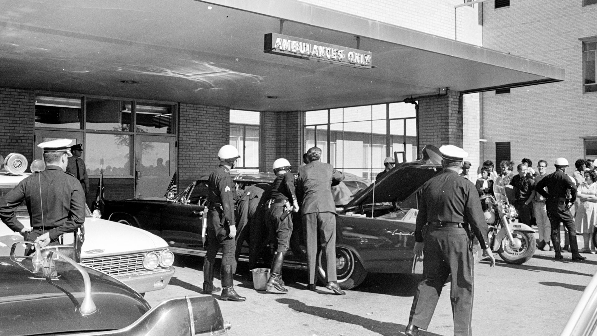 Police officers gather around the Presidential limousine (Lincoln-Mercury Continental) in front of an entrance to Parkland Hospital in Dallas, Texas, following the arrival of President John F. Kennedy.