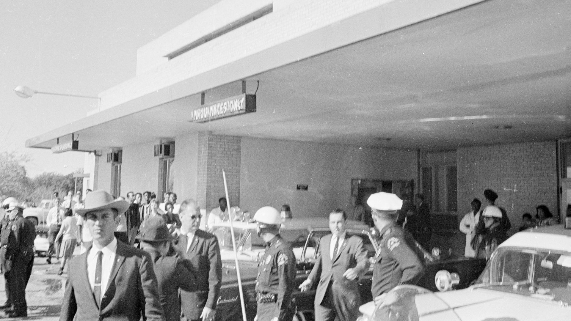Police officers and bystanders gather in front of an entrance to Parkland Hospital in Dallas, Texas, following the arrival of the Presidential limousine carrying President John F. Kennedy.