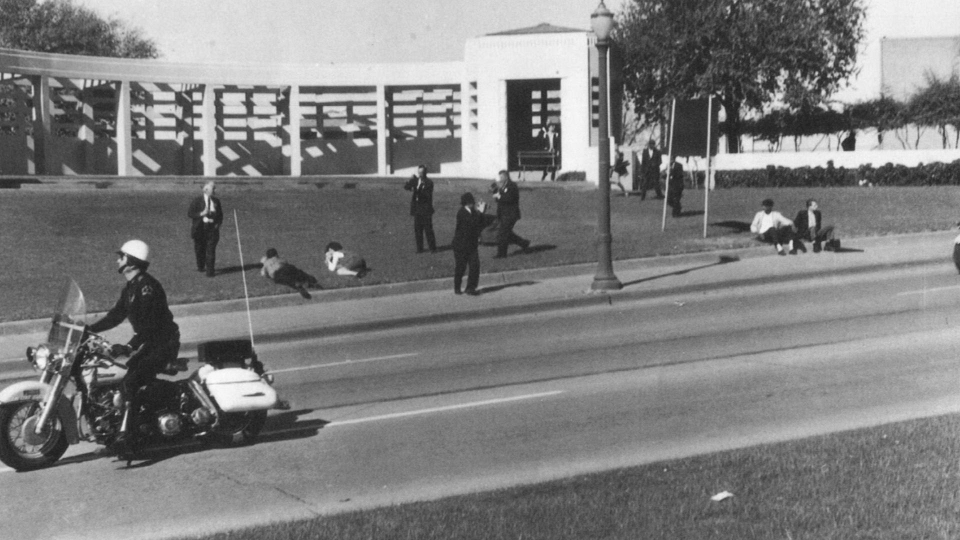 Assassination of JFK aftermath at Dealey Plaza; The Umbrella Man is sitting next to the road sign (the man on the right side)