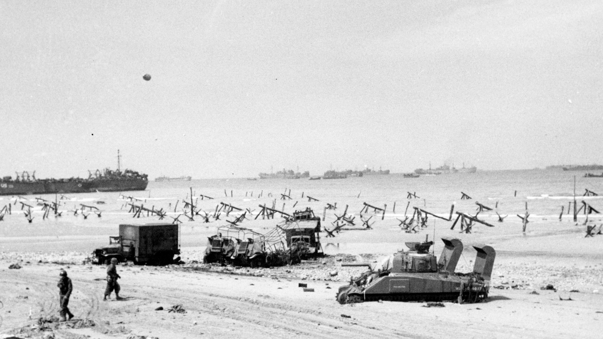 Normandy Invasion, June 1944: Scene on Omaha Beach on the afternoon of D-Day, 6 June 1944, showing casualties on the beach, a bogged-down M4 Sherman tank, several wrecked trucks and German anti-landing obstructions. A LST is beached in the left distance and invasion shipping is off shore.
