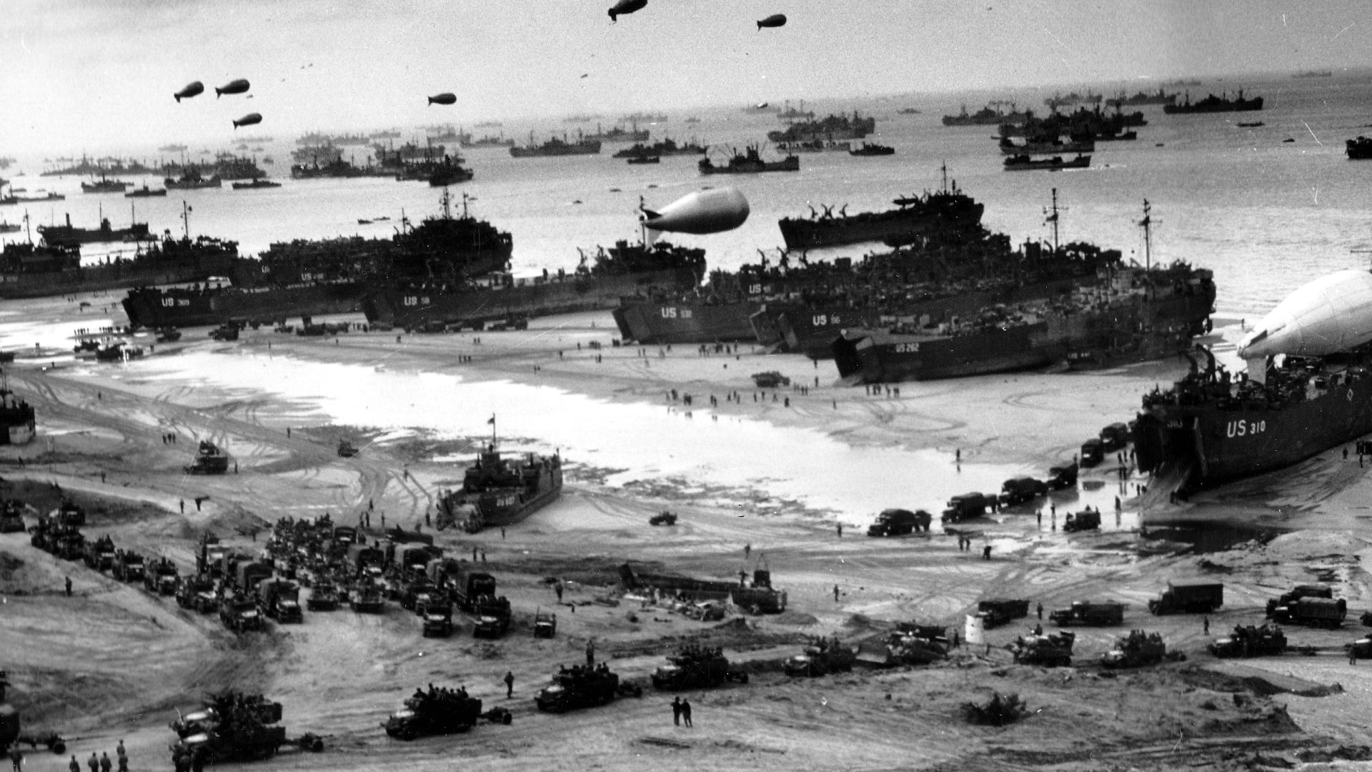 Normandy Invasion, June 1944. Landing ships putting cargo ashore on one of the invasion beaches, at low tide during the first days of the operation, June 1944. Among identifiable ships present are USS LST‑532 (in the center of the view); USS LST‑262 (third LST from right); USS LST‑310 (second LST from right); USS LST‑533 (partially visible at far right); and USS LST‑524. Note the barrage balloons overhead and Army
