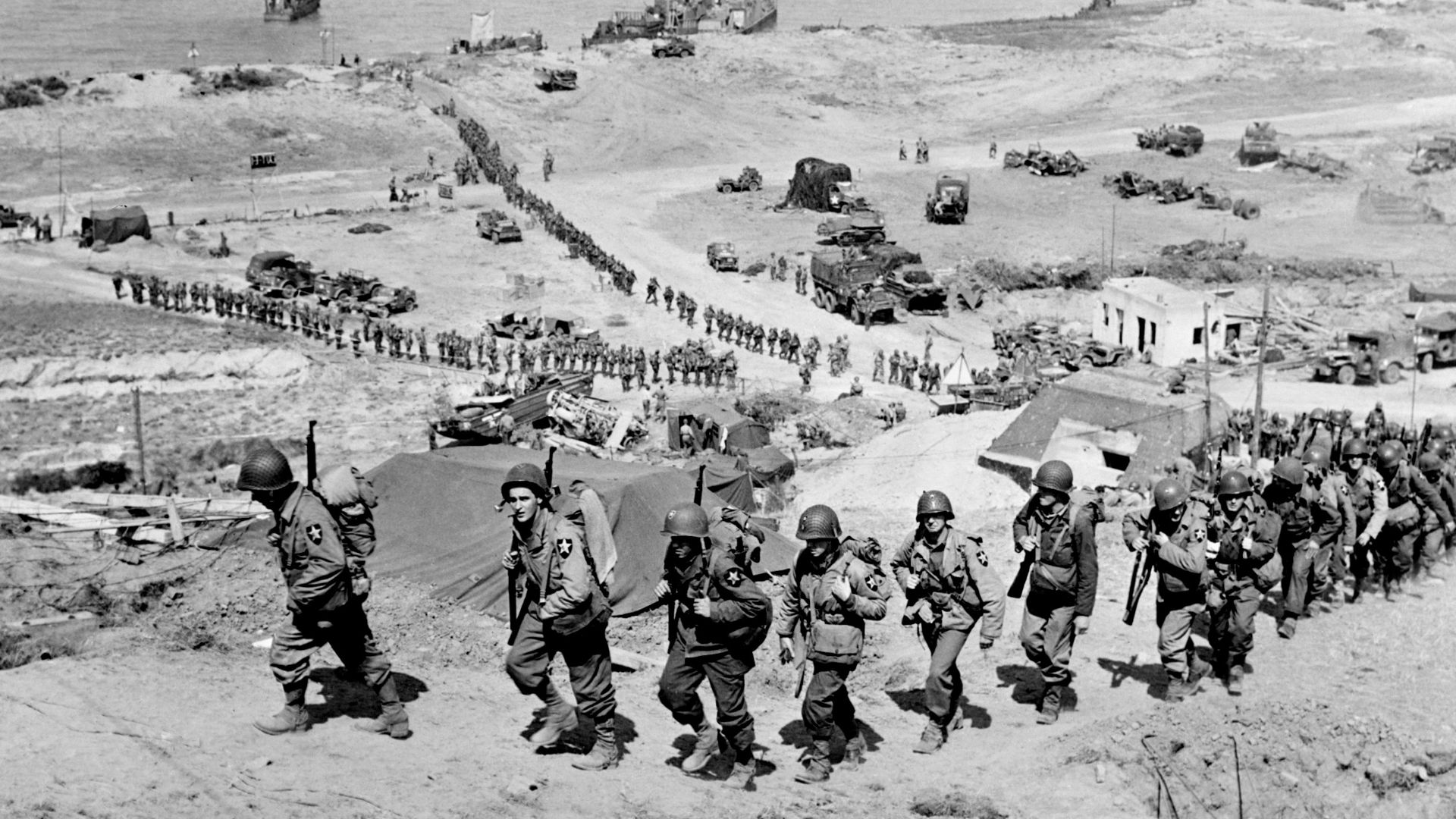 Troops of the US Army 2nd Infantry Division march up the bluff at the E-1 draw in the Easy Red sector of Omaha Beach, Normandy, France on D+1, June 7, 1944. They are going past the German bunker, Widerstandsnest 65 (WN 65), that defended the route up the Ruquet Valley to Saint-Laurent-sur-Mer.