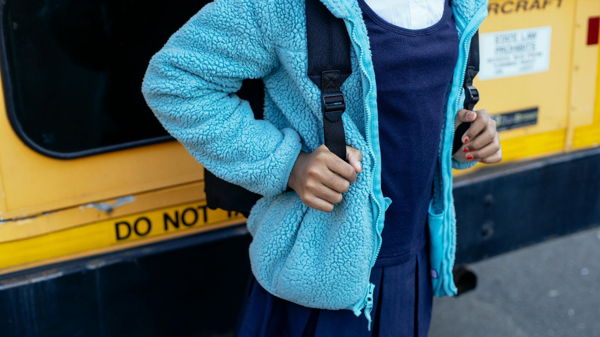A schoolgirl in a blue jacket standing near a yellow school bus with a backpack.