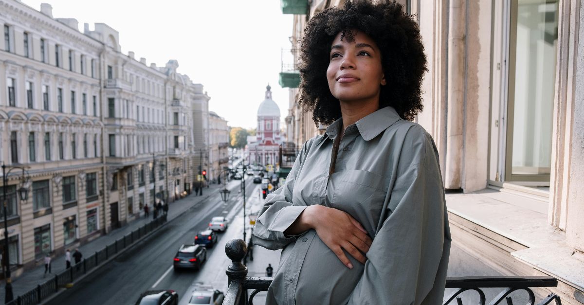 A pregnant woman stands on a balcony overlooking a bustling city street, radiating tranquility.