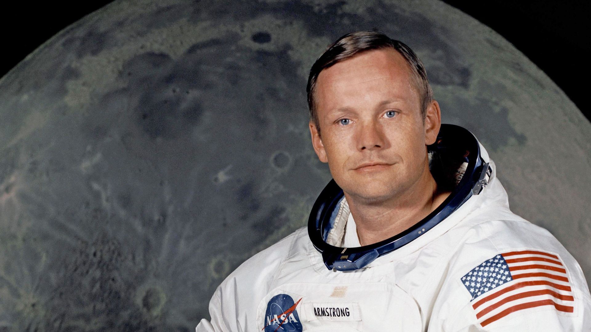 Portrait of Astronaut Neil A. Armstrong, commander of the Apollo 11 Lunar Landing mission in his space suit, with his helmet on the table in front of him. Behind him is a large photograph of the lunar surface.