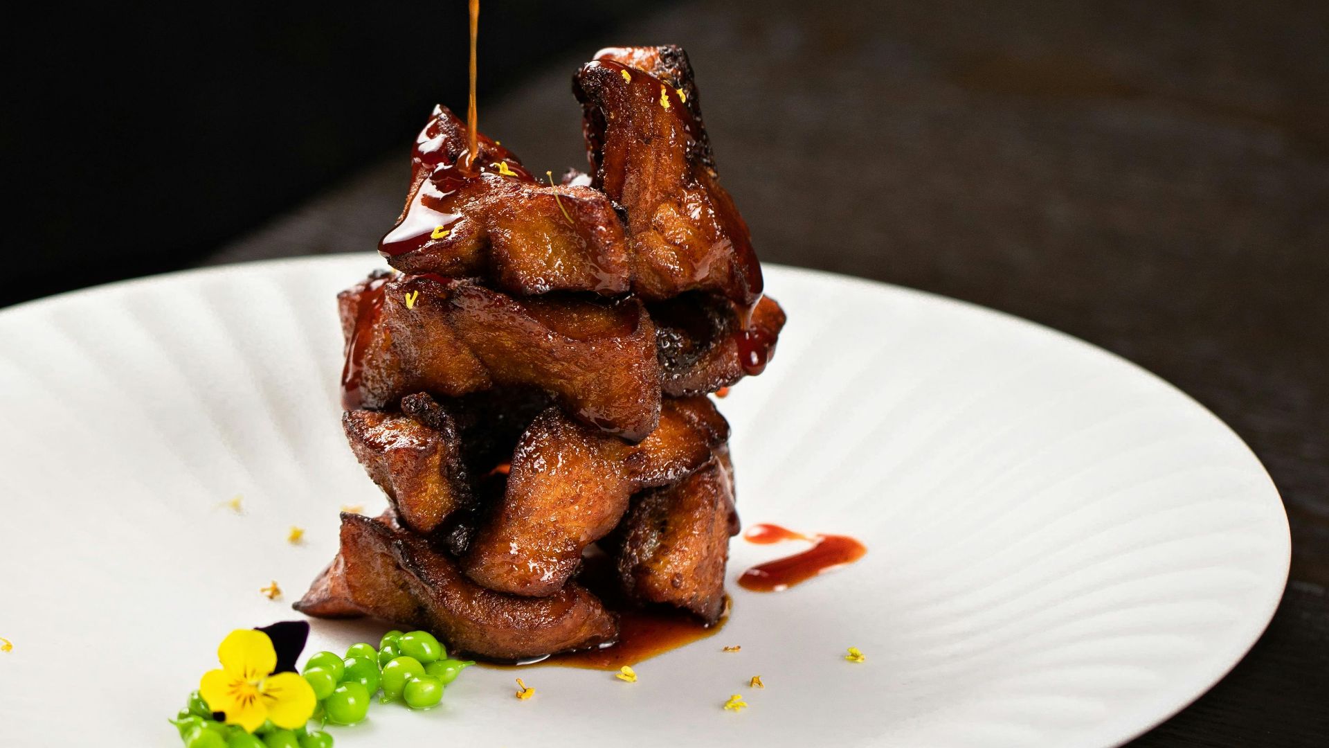 Close-up of a hand pouring sauce over artistically arranged glazed meat on a plate.
