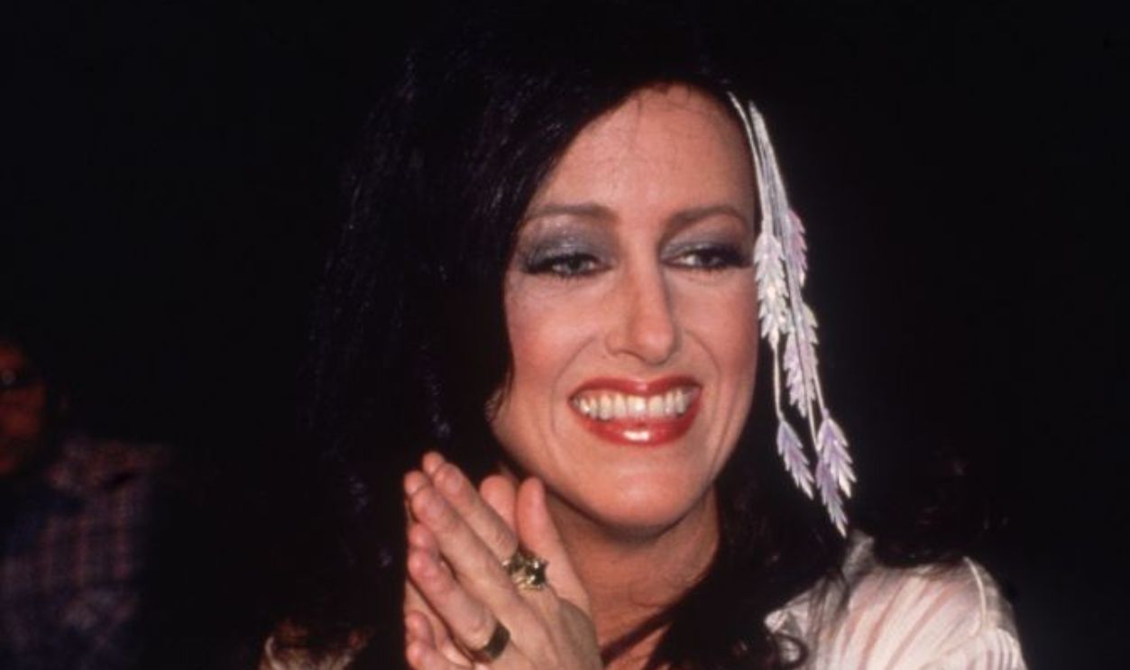 circa 1979: American singer Grace Slick smiling, holding her hands clasped together. She is wearing a white outfit, a white belt, and a gold lion's head belt buckle.