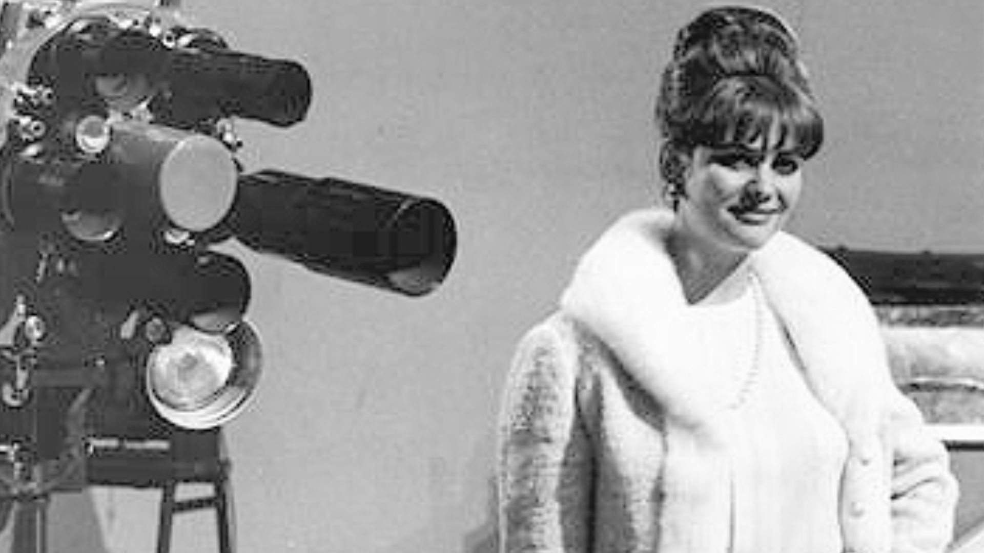 Studio-plateau avec à gauche, une caméra de télévision et à droite, une jeune femme brune avec chignon et frange, souriante, se tenant debout, bras gauche sur la hanche et tenant de l'autre bras, un sac à main de cuir foncé.