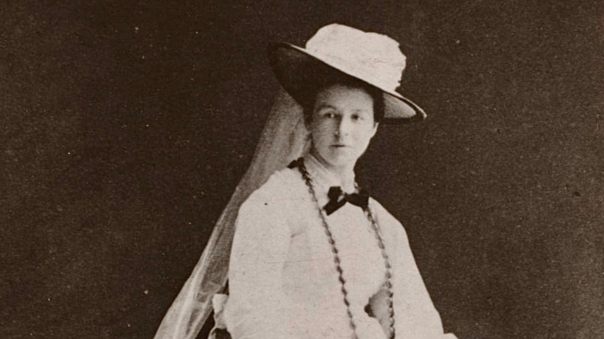 C. 1870s. Wearing a long, light colored gown with a hopped skirt, a brimmed hat with a train and a beaded necklace. She’s resting her arm on a pedestal.