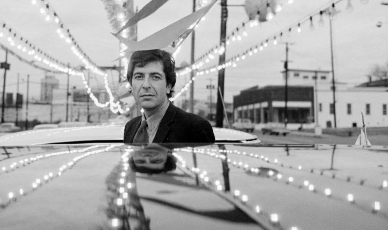 TENNESSEE, NASHVILLE - 1968: Leonard Cohen by a car at a car dealer ship and over his head are flags and lights.