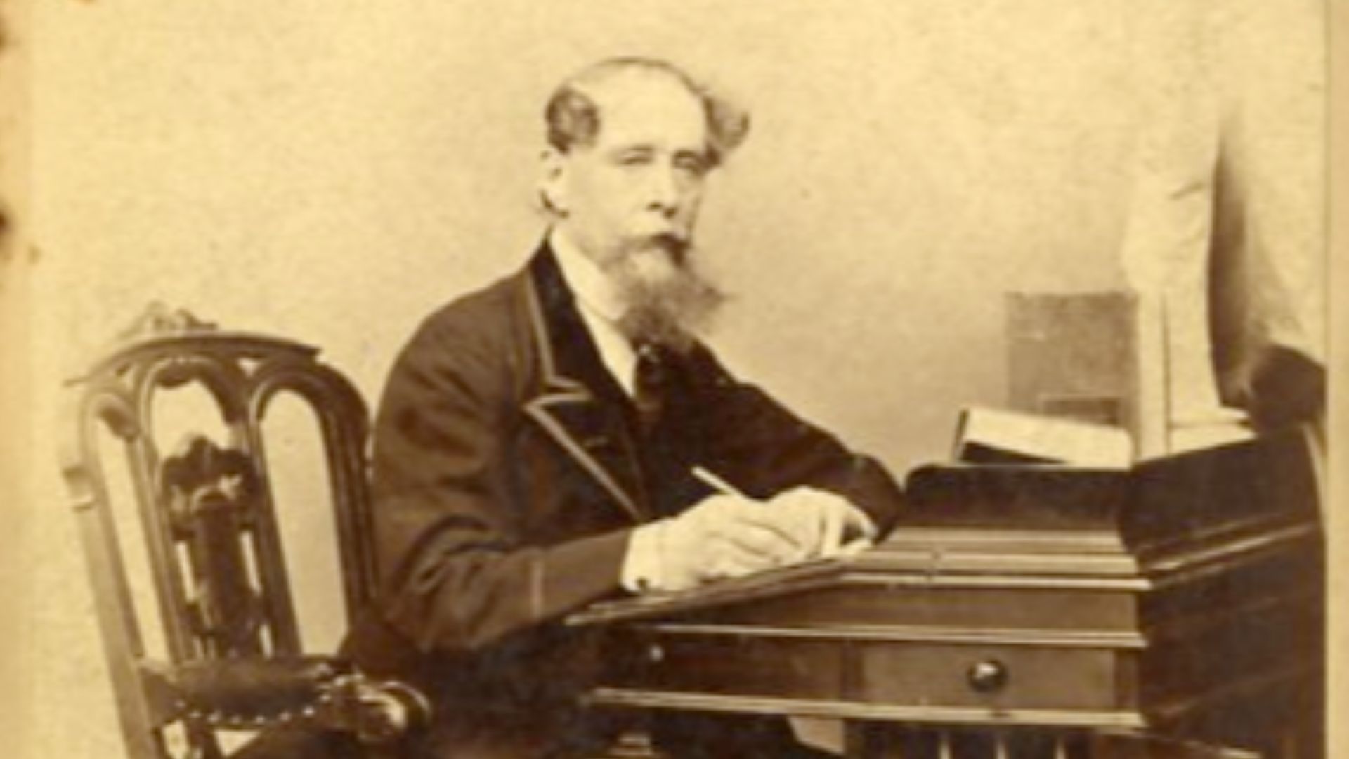 Charles Dickens sits at his desk with his pen in hand surrounded by books and folios