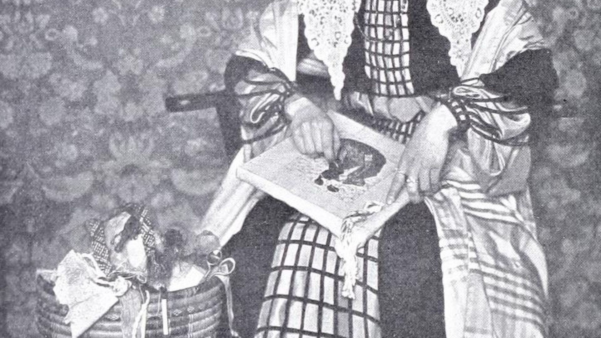 A photograph of a woman sitting doing embroidery in front of a patterned wallpaper.