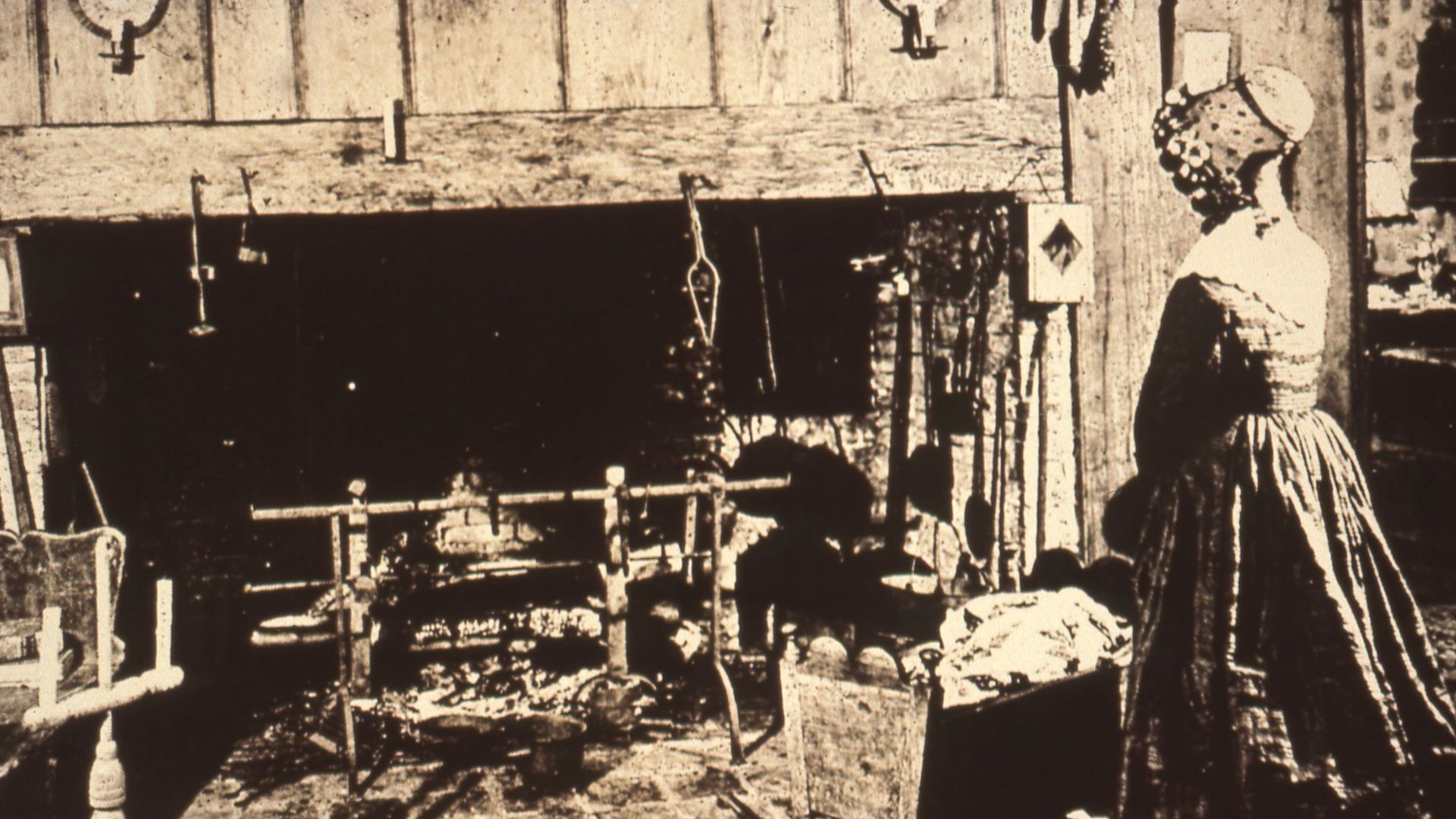 A woman wearing a bonnet stands facing a large hearth, a child's cradle nearby. Large andirons are in the fireplace, and various cooking tools hang within reach. A striped rug lies on the floor,