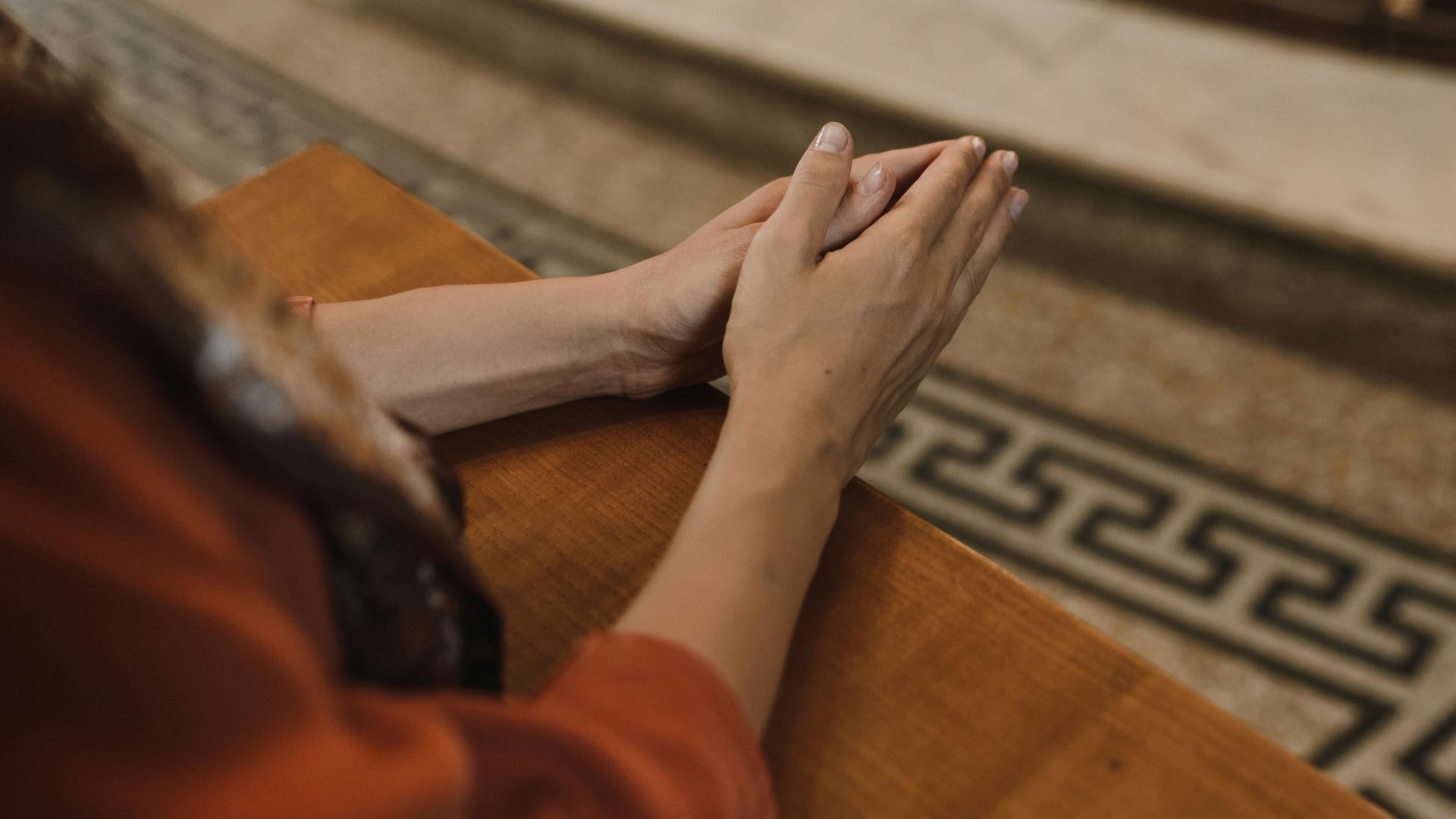 A close-up image of hands together in prayer on a wooden surface in an indoor setting.