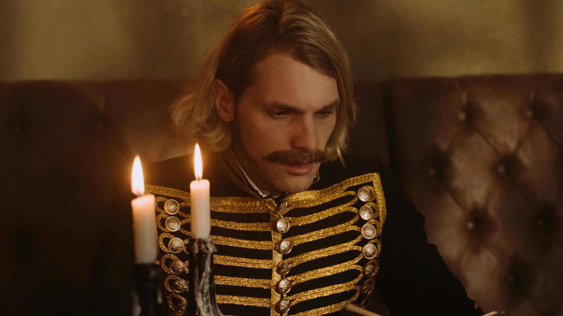 A man with blond hair reads a book by candlelight, dressed in a historical uniform in an antique room.