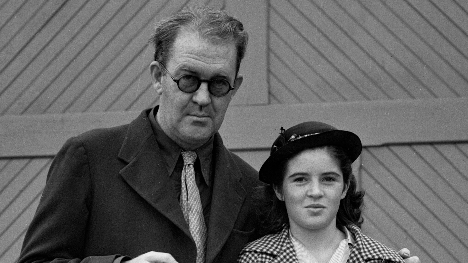 John Ford and his daughter Barbara Ford after returning from Hawaii. They are outside, and John holds a pipe while Barbara holds a ukulele and a purse. Text from newspaper caption: John Ford, Fox director, returned on the Matson liner Malolo with his family yesterday after six weeks’ absence to Hawaii, and ready to start work on The Message to Garcia. With him were Mrs. Ford, their daughter, Barbara, and their son, Pat, the latter having sailed with his father to Hawaii from here in their ketch Aranar. Ford is with his daughter.