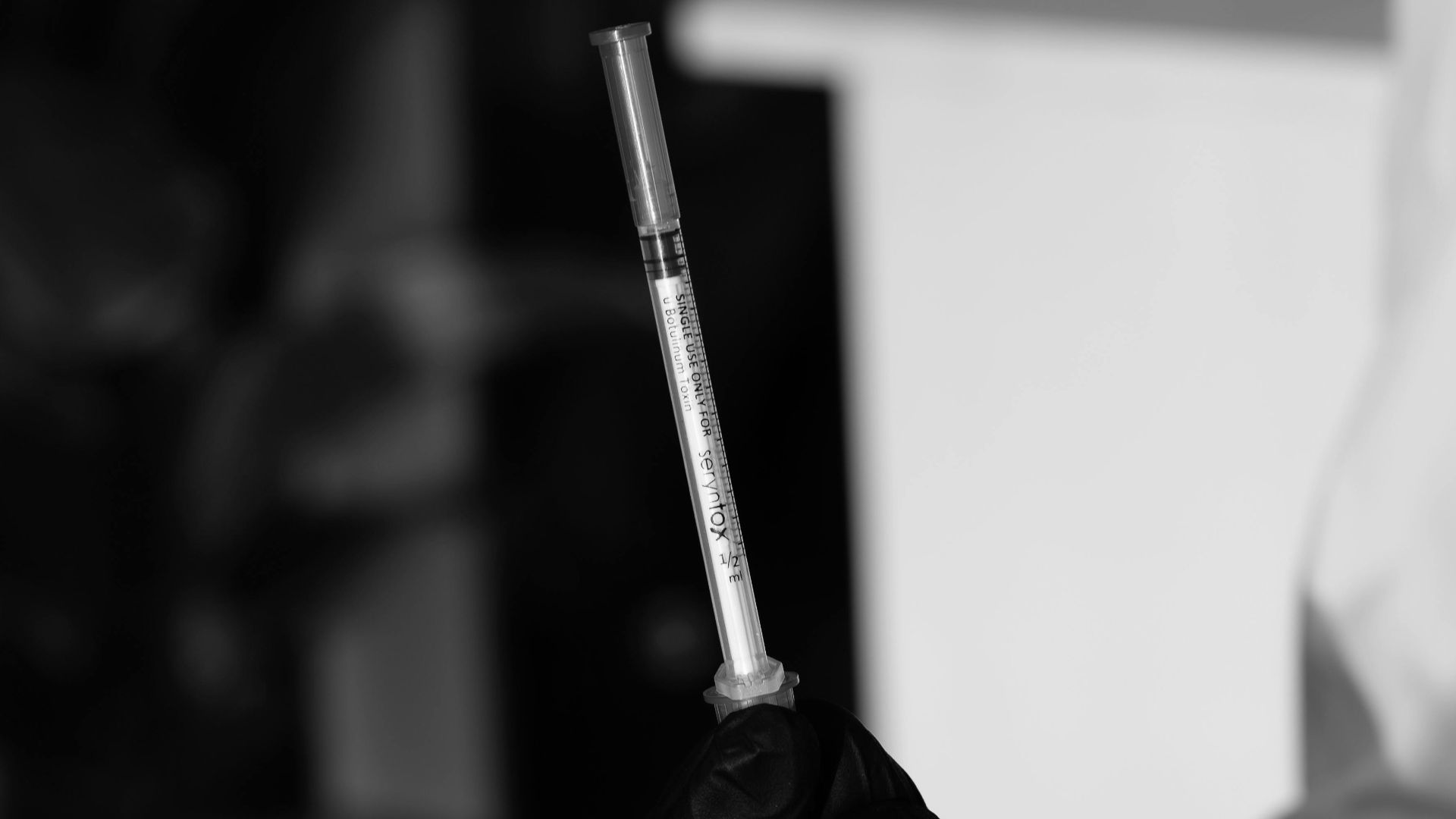 Close-up black and white image of a syringe held by a gloved hand, emphasizing medical precision.