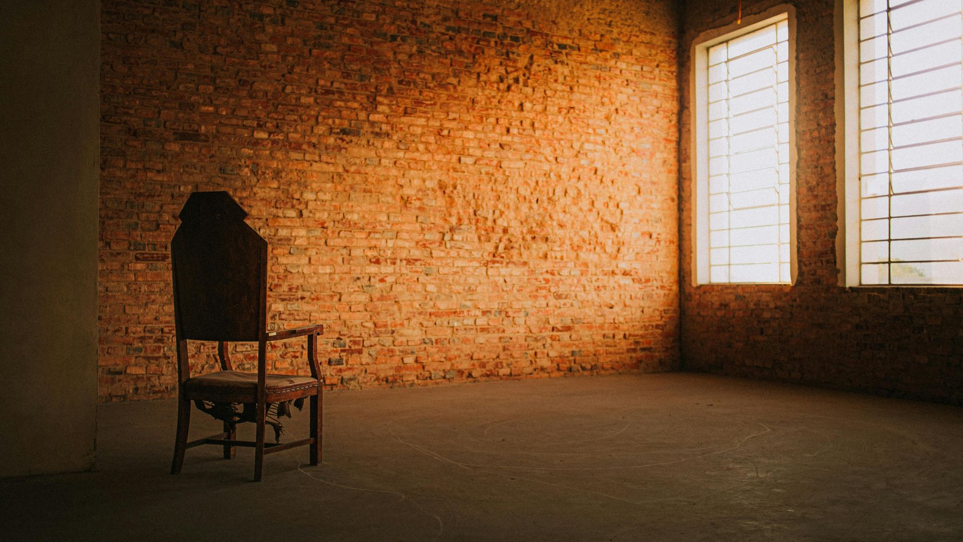 A minimalistic room features a solitary chair against a textured brick wall, creating a moody atmosphere.