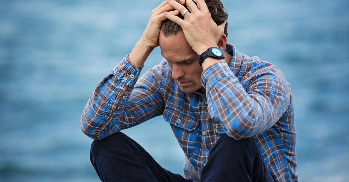 A man in a plaid shirt sits by the water looking distressed, symbolizing stress.