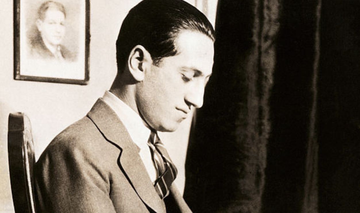 (Original Caption) George Gershwin (1898-1937), American composer, is shown seated at a piano. He holds a cigar and sheet music in his lap. 