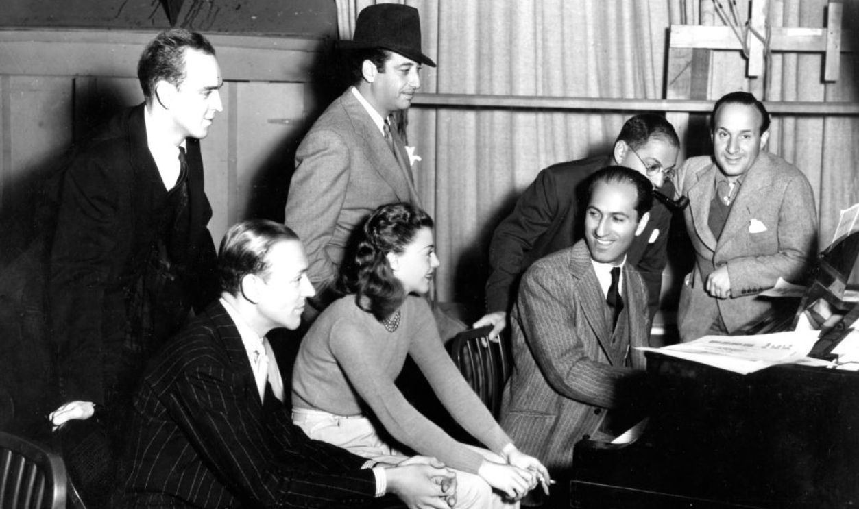 On the set of the film 'Shall We Dance,' George Gershwin plays piano with, from left, dance director Hermes Pan, actors Fred Astaire and Ginger Rogers, film director Mark Sandrich, Ira Gershwin, and musical director Nathaniel Shilkret, Hollywood, California, 1936. 