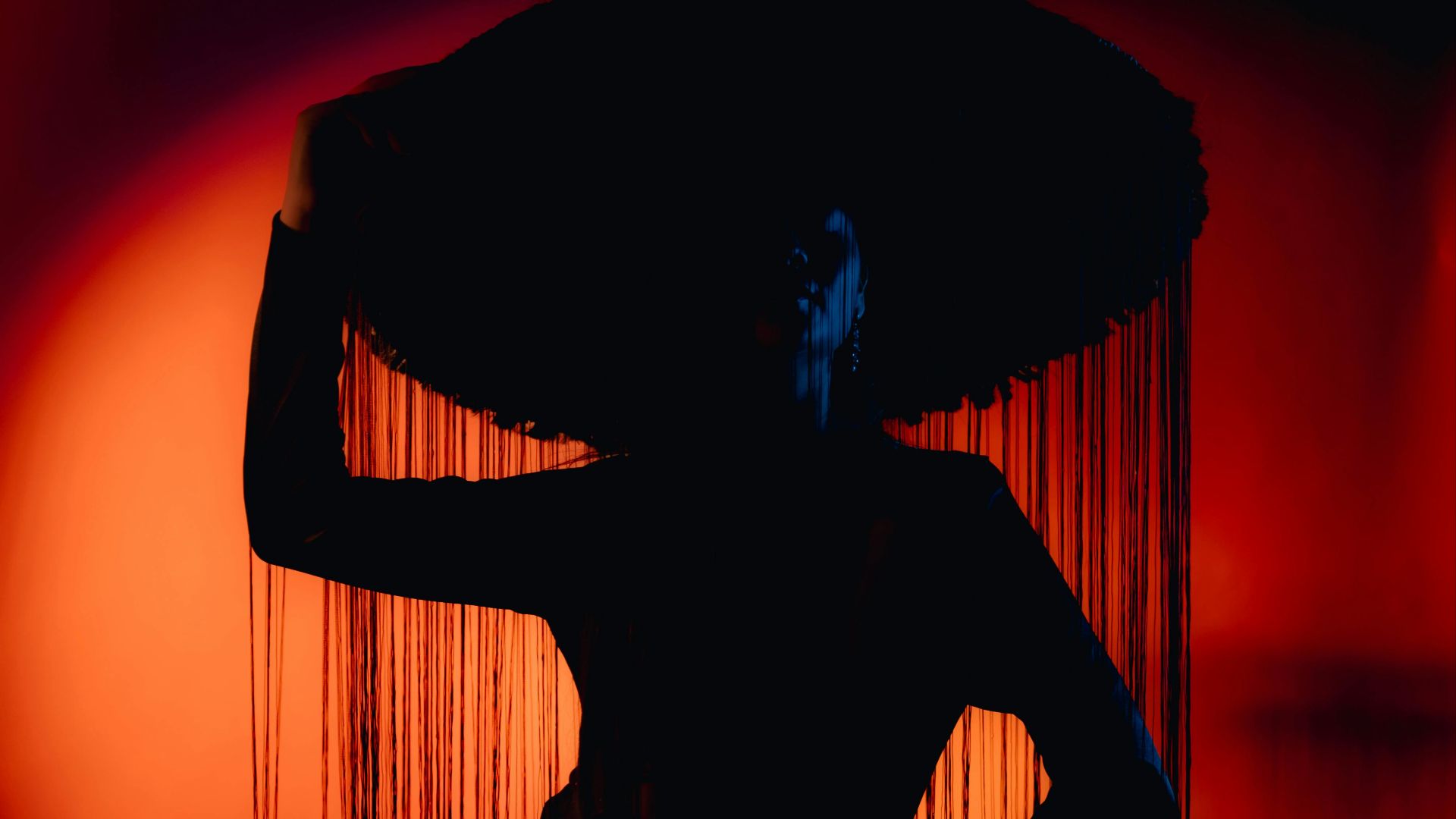 Silhouette of a stylish figure in a dramatic outfit and fringed hat against an orange backdrop.