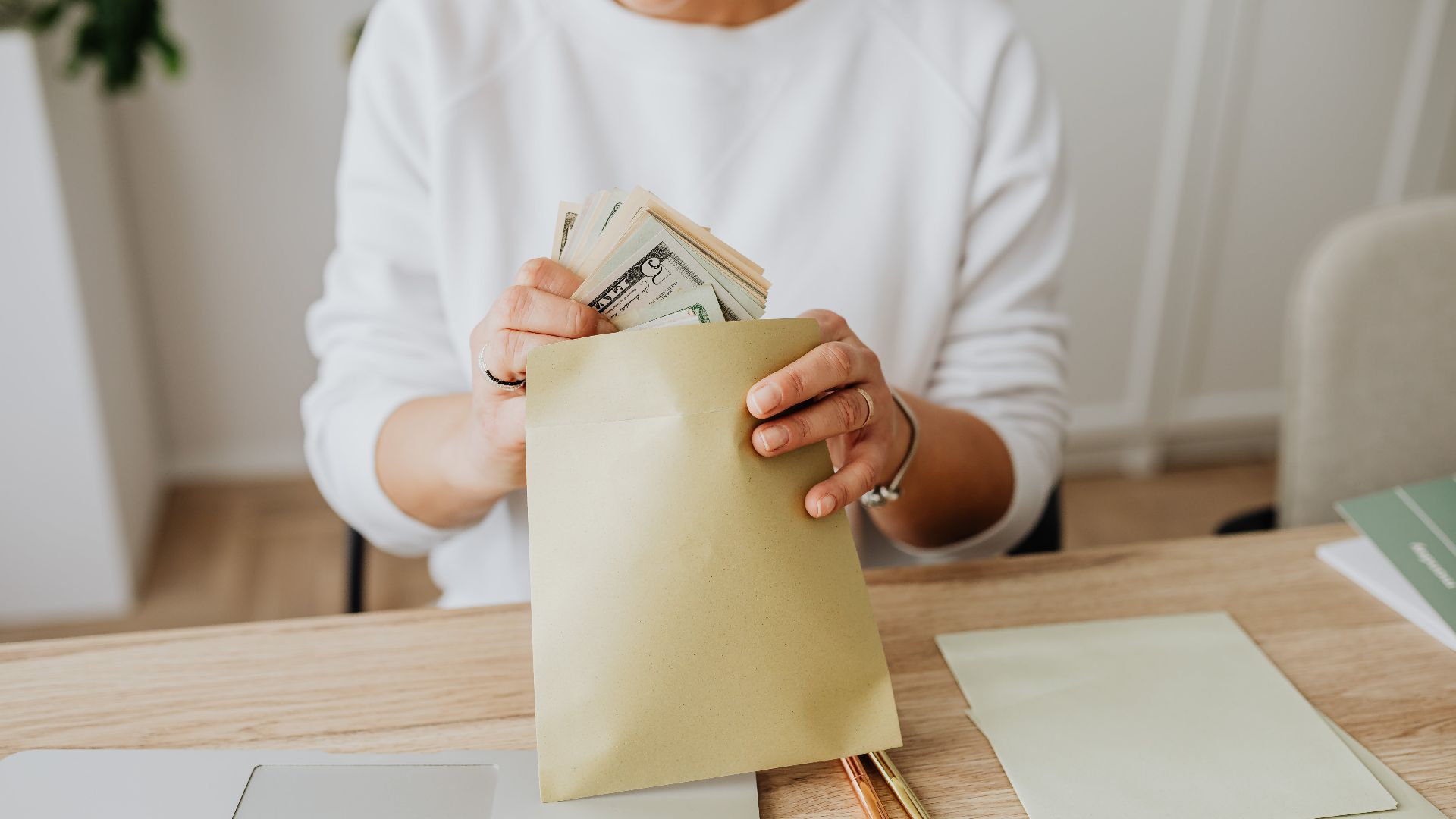 Close-up of person holding envelopes with cash at a wooden desk indoors.