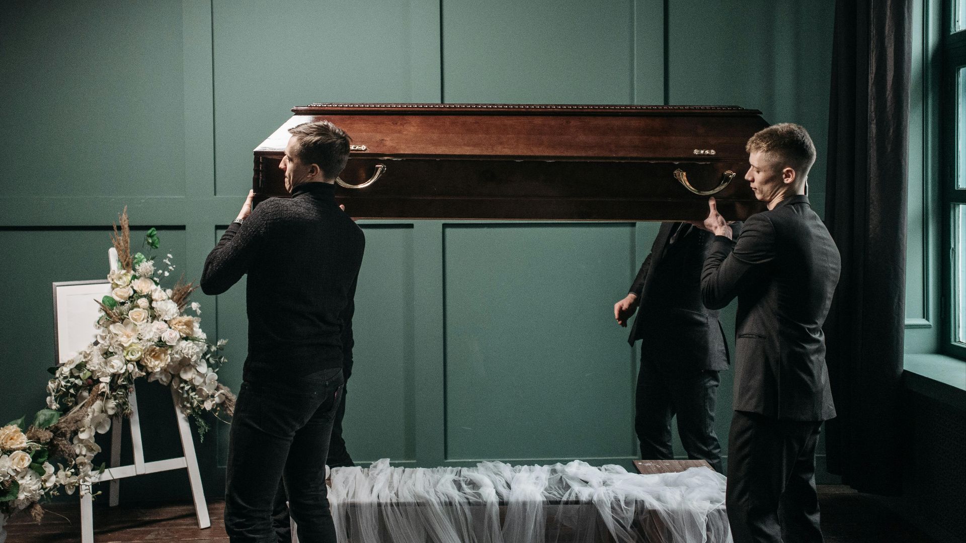 Pallbearers carry a wooden coffin at an indoors funeral service, surrounded by floral tributes.