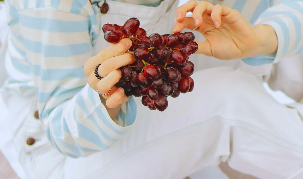 Woman with Curly Hair Sitting on a Picnic Blanket while Looking at the Grapes she is Holding