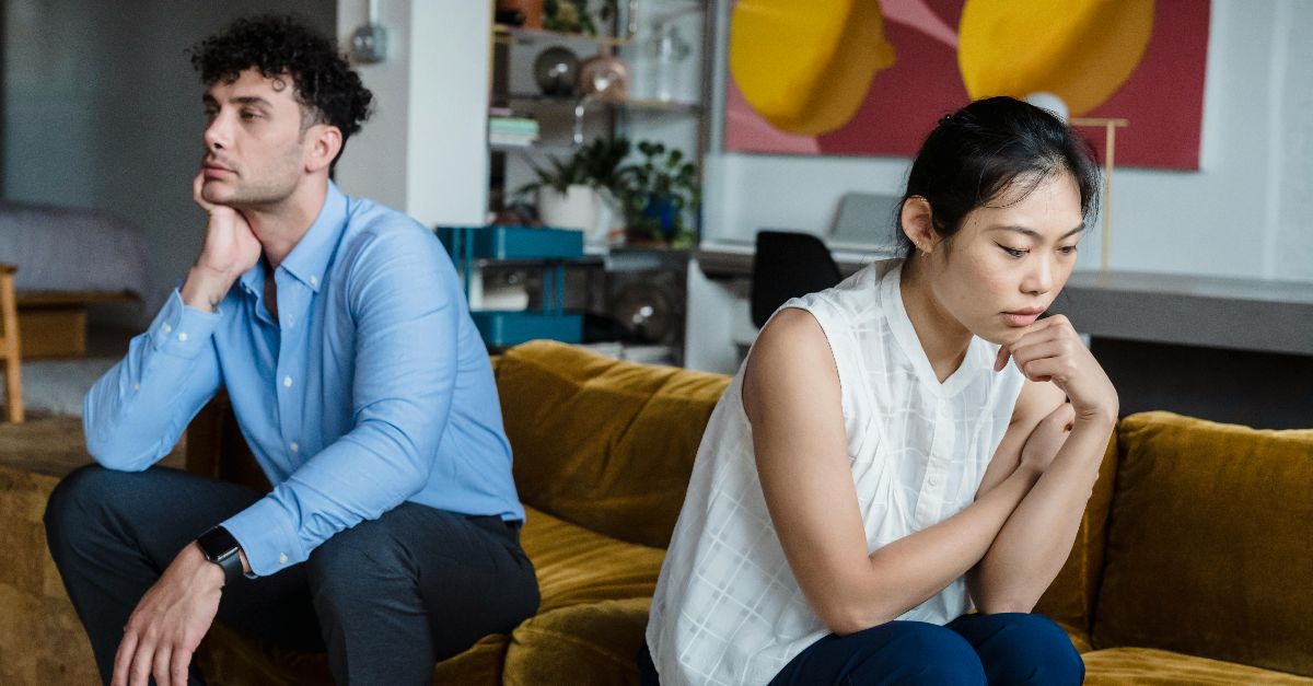 A couple sits indoors on a sofa, both appearing thoughtful and contemplative.