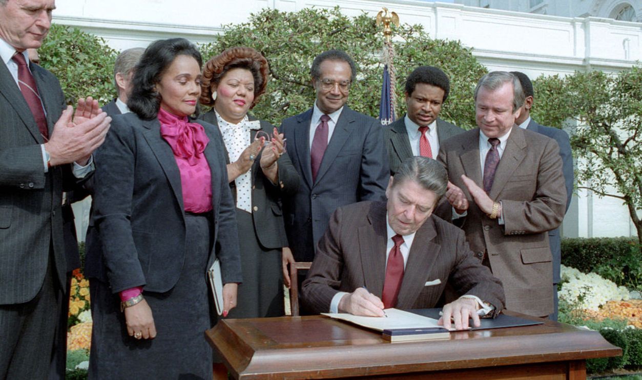 American Civil Rights activist Coretta Scott King (1927 - 2006) (second left) and others watch as US President Ronald Reagan (1911 - 2004) signs the Martin Luther King Holiday Legislation during a ceremony in the White House's Rose Garden, Washington DC, November 11, 1983. Among those pictured are, from left, Vice President George HW Bush (1924 - 2018), King, US Representative Katie Hall (1939 - 2012), Secretary of Housing & Urban Development Samuel Pierce (1922 - 2000), unidentified, and Senator Howard Baker (1925 - 2014). 