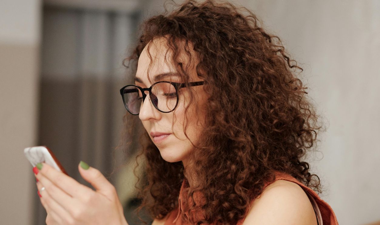 Woman with curly hair and eyeglasses using smartphone at cafe table, daytime