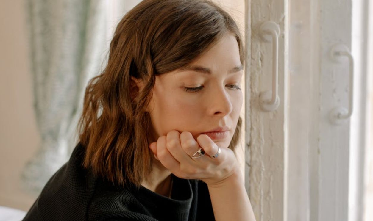 Young woman looking thoughtful while sitting by a window indoors. Natural lighting and contemplative mood.