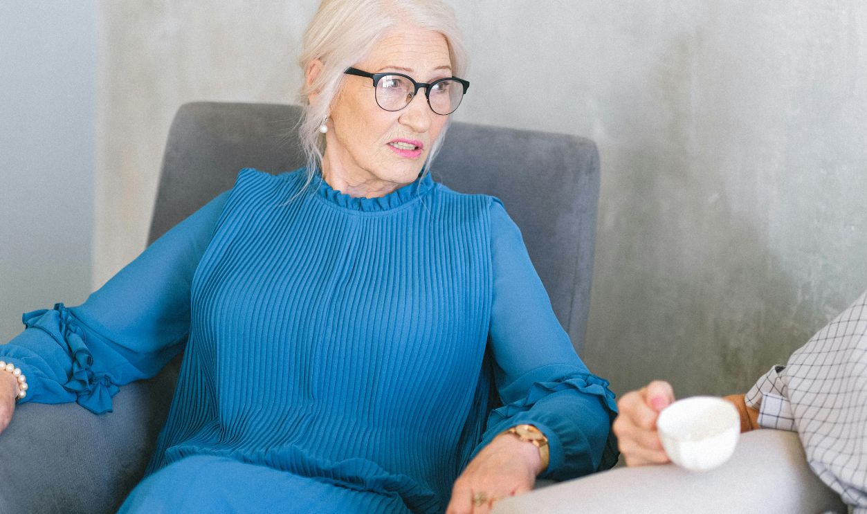 Senior woman in a stylish blue dress conversing in a serene indoor setting, exuding elegance and comfort.