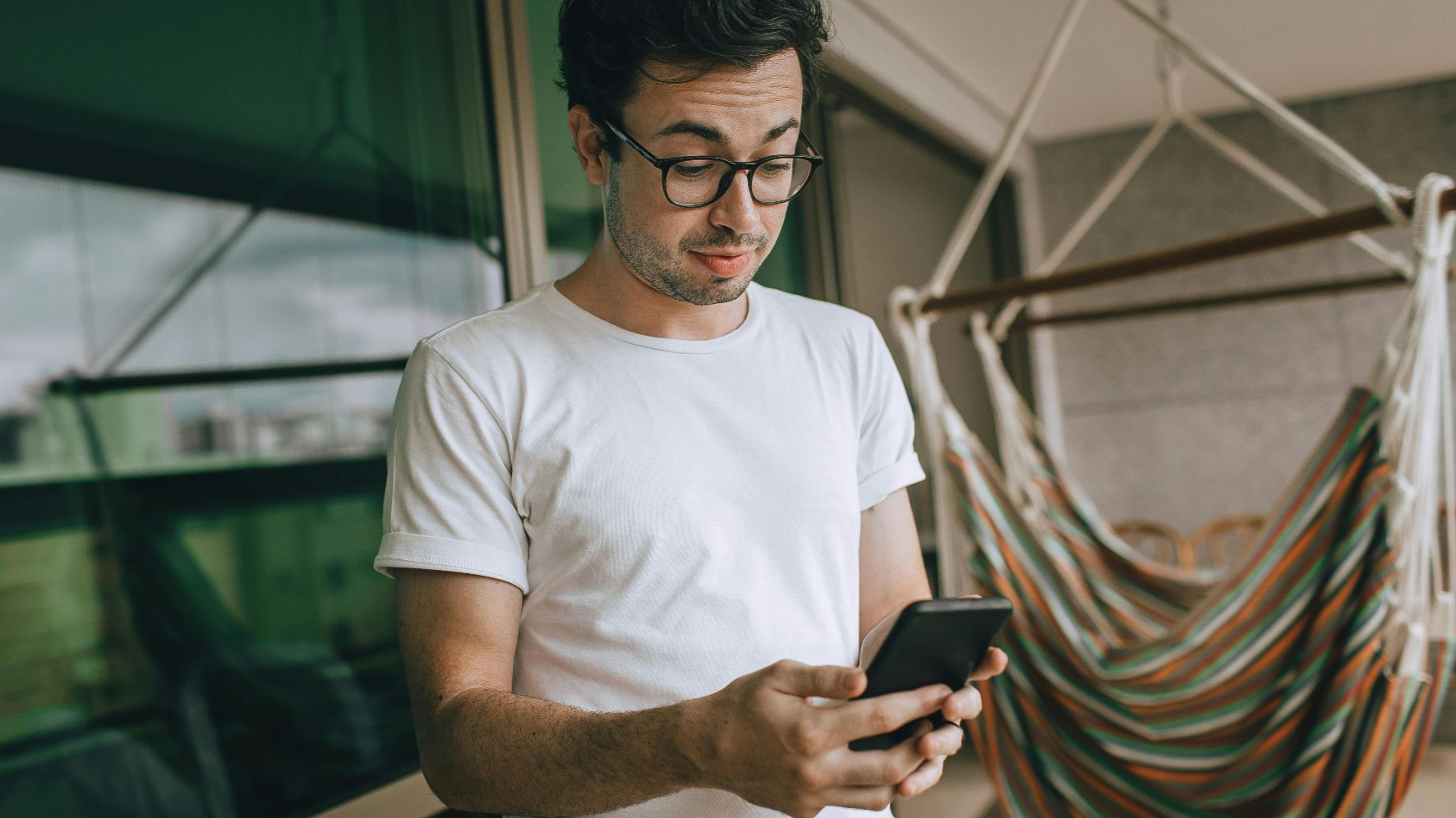 Young man in glasses using smartphone indoors with striped hammock.