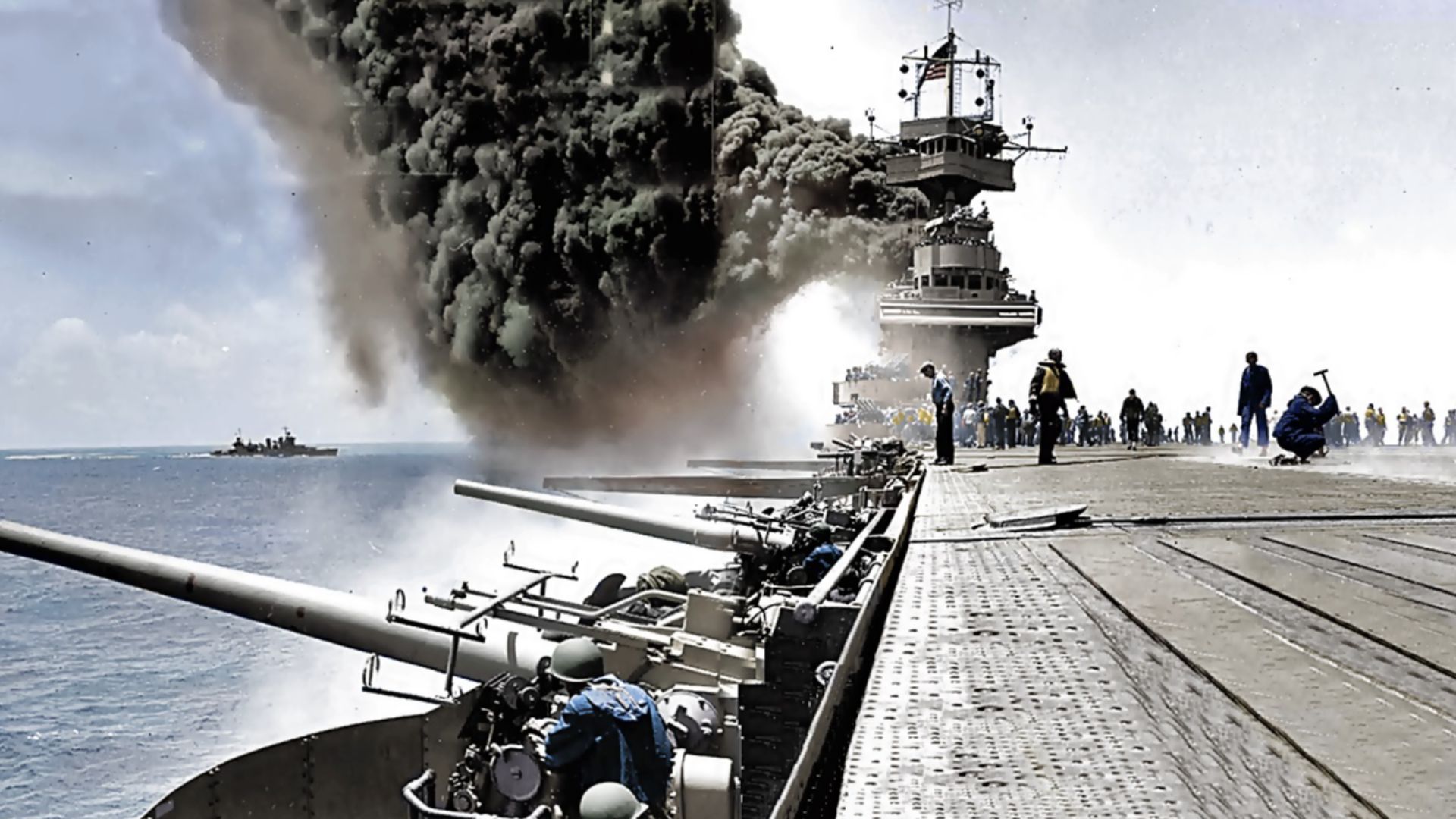 Scene on board USS Yorktown (CV-5), during the Battle of Midway, 4 June 1942, shortly after she was hit by three Japanese bombs on the same day, what would cause the sinking of the aircraft carrier on 7 June 1942, in which 141 men were killed. The Battle of Midway, however, was a decisive naval and air battle for the US Navy and widely considered a turning point in the Pacific War. Dense smoke is from fires in her uptakes, caused by a bomb that punctured them and knocked out her boilers. The engineer with the hammer at the right side is most likely covering a bomb entry hole in the forward elevator. Note arresting gear cables and forward palisade elements on the flight deck; CXAM radar antenna, large national ensign and YE homing beacon antenna atop the foremast; 5