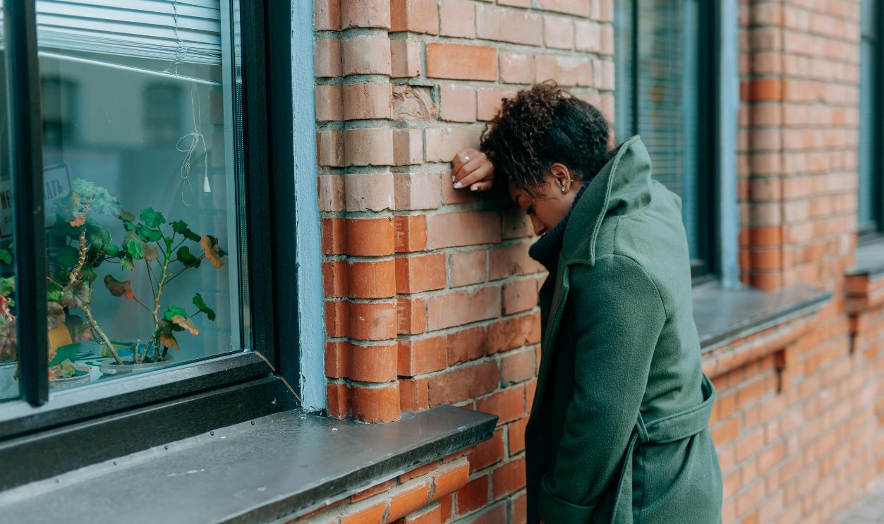 A woman in a green coat leans against a brick wall, reflecting deeply outdoors