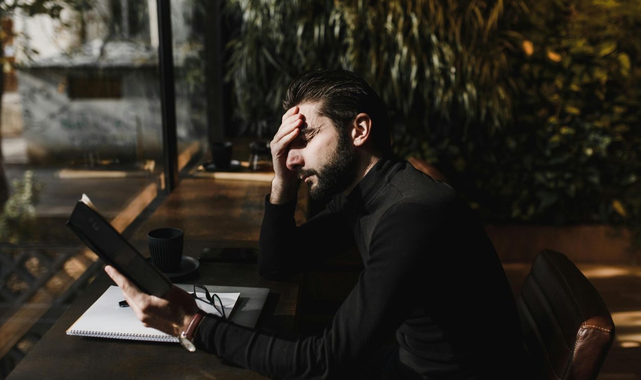 A stressed man with hand on forehead reads in a sunlit room, illustrating concentration and pressure