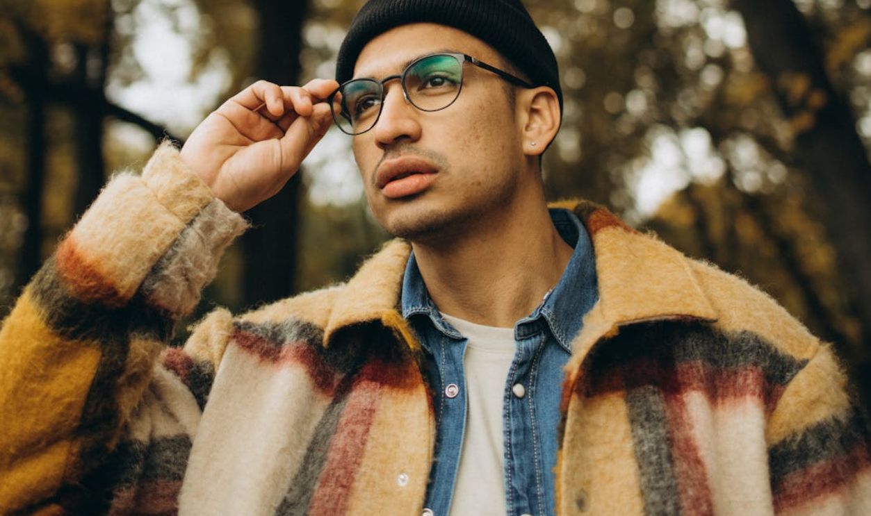 A fashionable man with eyewear and a checkered coat thoughtfully posing in a serene fall setting