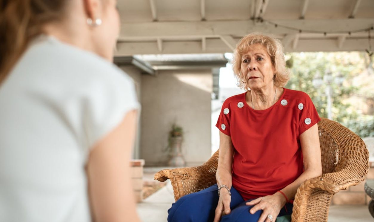 Senior woman in red shirt conversing indoors with younger woman. Daytime scene