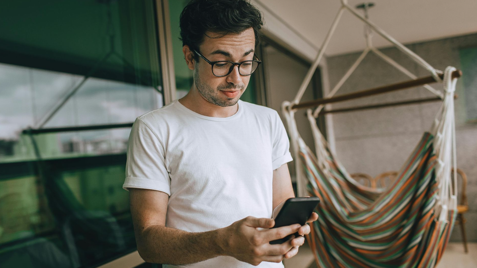 Young man in glasses using smartphone indoors with striped hammock